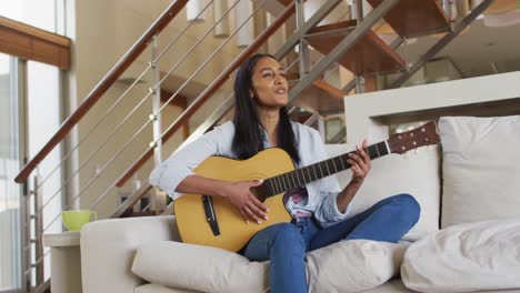 mujer de raza mixta en el sofá en casa tocando la guitarra y cantando