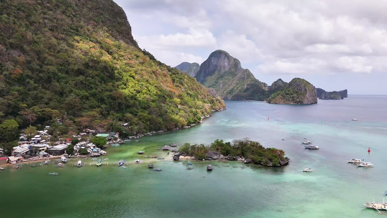 Tropical coastline in El Nido, Palawan with turquoise waters, scattered boats, small village, and lush limestone cliffs under partly cloudy skies in a scenic, tranquil island setting