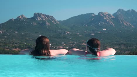 Dos-Chicas-Jóvenes-Se-Relajan-En-Una-Piscina-Infinita-Mirando-Un-Pintoresco-Paisaje-Montañoso-Sardo