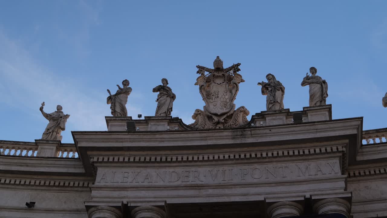4K shot of stone statues and papal crest above the Alexander VII inscription on the colonnade of St. Peter’s Square, Vatican City, Rome, with clear blue sky and historic detail