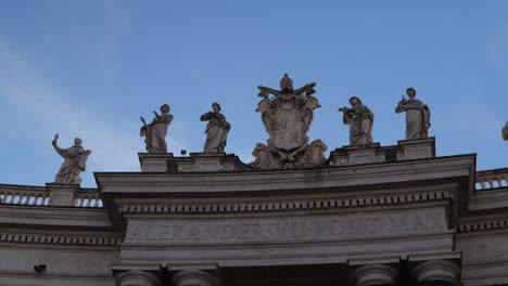 4k shot of stone statues and papal crest above the alexander vii inscription on the colonnade of st. peter’s square, vatican city, rome, with clear blue sky and historic detail