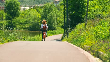 Girl-riding-bicycle-on-empty-road-of-tranquil-park-with-green-lush-vegetation-on-a-sunny-day