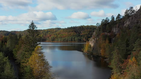 Aerial-drone-view-over-trees,-a-calm-lake-and-cliffs,-in-middle-of-hills-and-foliage-forest,-sunny,-autumn-morning,-in-South-Norway