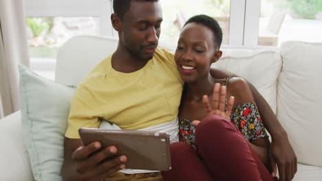 Happy-african-american-couple-sitting-on-sofa-embracing-and-talking-looking-at-tablet