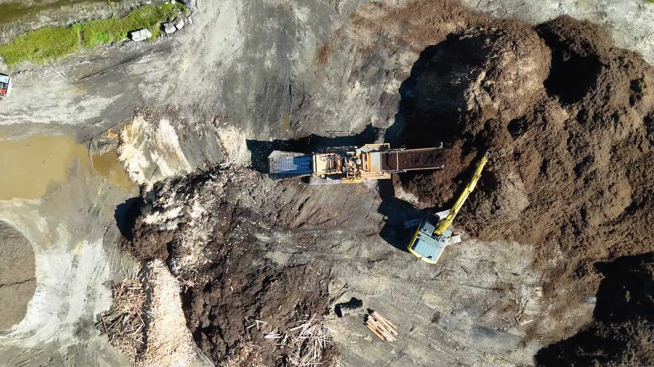 Drone footage captures an excavator moving mulch in an industrial setting under bright daylight