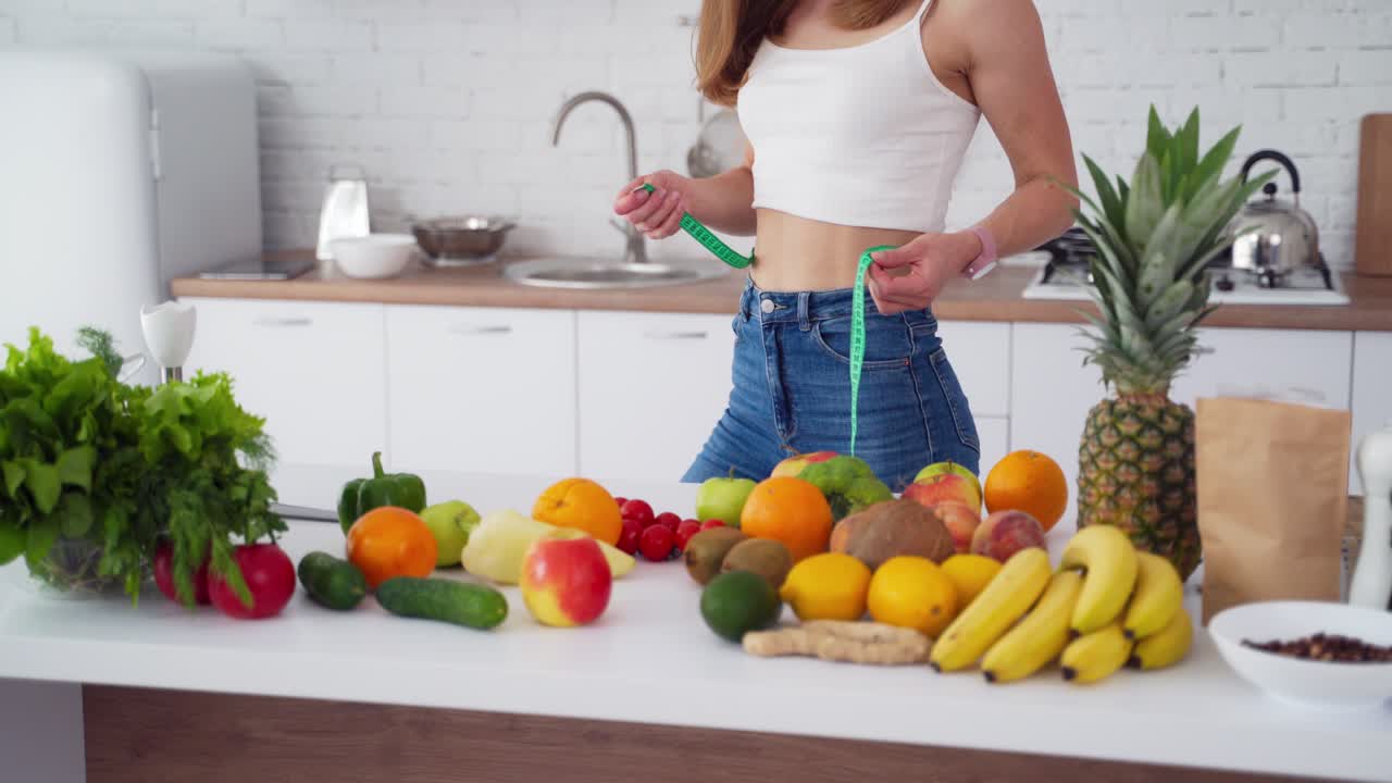 Sporty young woman in light kitchen. Attractive sporty woman standing on kitchen with centimeter while measuring her waist