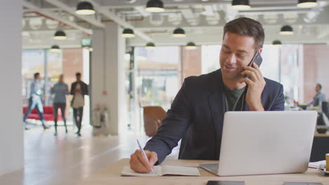 Businessman-Sitting-At-Desk-On-Phone-Call-In-Modern-Open-Plan-Office-With-Colleagues-In-Background