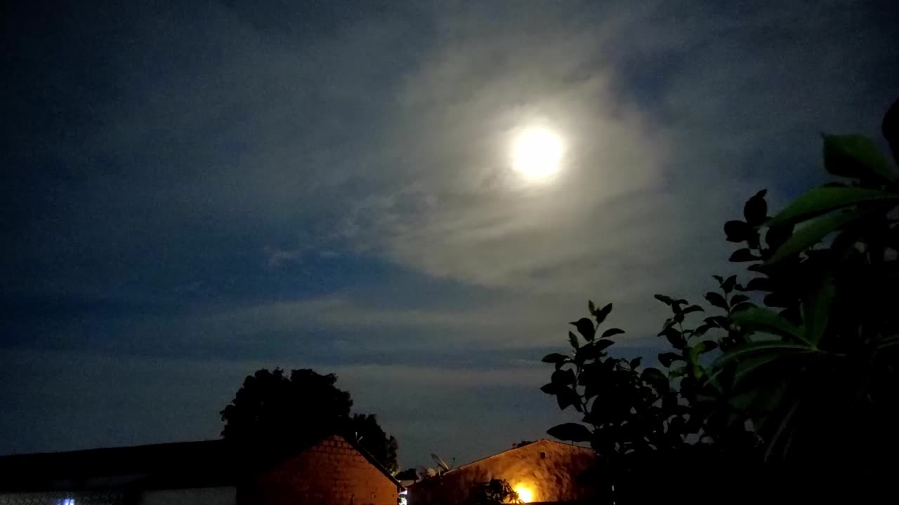 Time-lapse do movimento da lua da esquerda para a direita com telhados de casas e plantas visíveis em uma noite ligeiramente nublada.