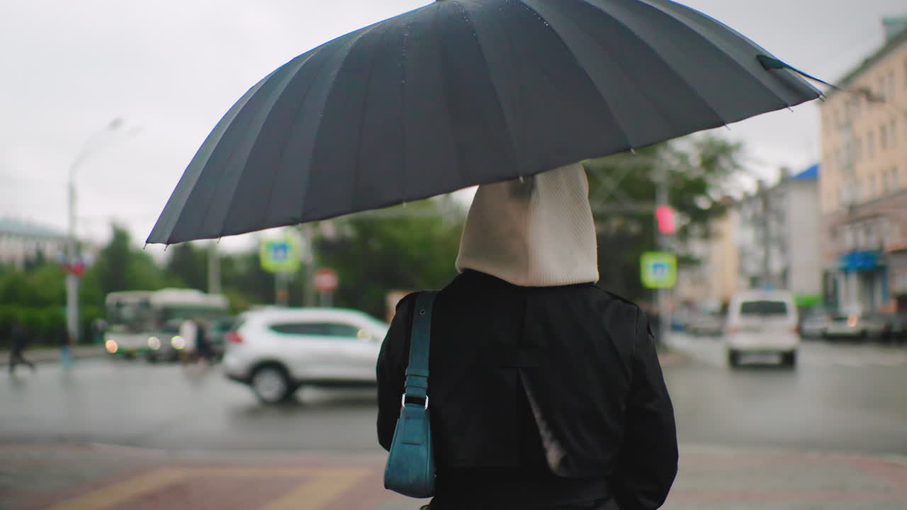 우산을 쓰고 빗오는 도시 거리 횡단보도를 걷는 후드 재킷을 입은 소녀, 흐릿한 보행자, 자동차, 건물이 배경에 있고 흐린 날씨 분위기의 도시 라이프스타일 장면이에요.