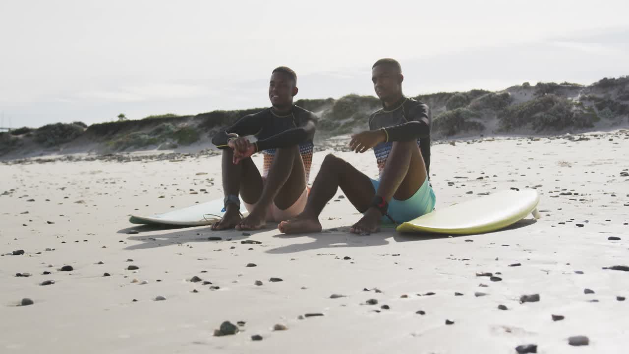 felices hermanos gemelos adolescentes afroamericanos sentados junto a las tablas de surf en una playa hablando