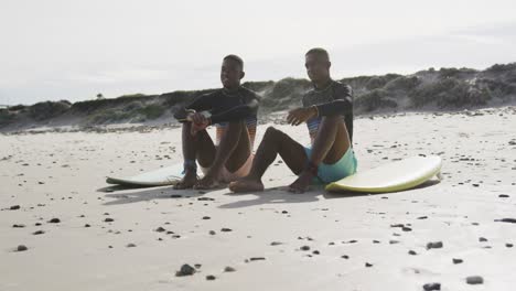 happy african american teenage twin brothers sitting by surfboards on a beach talking