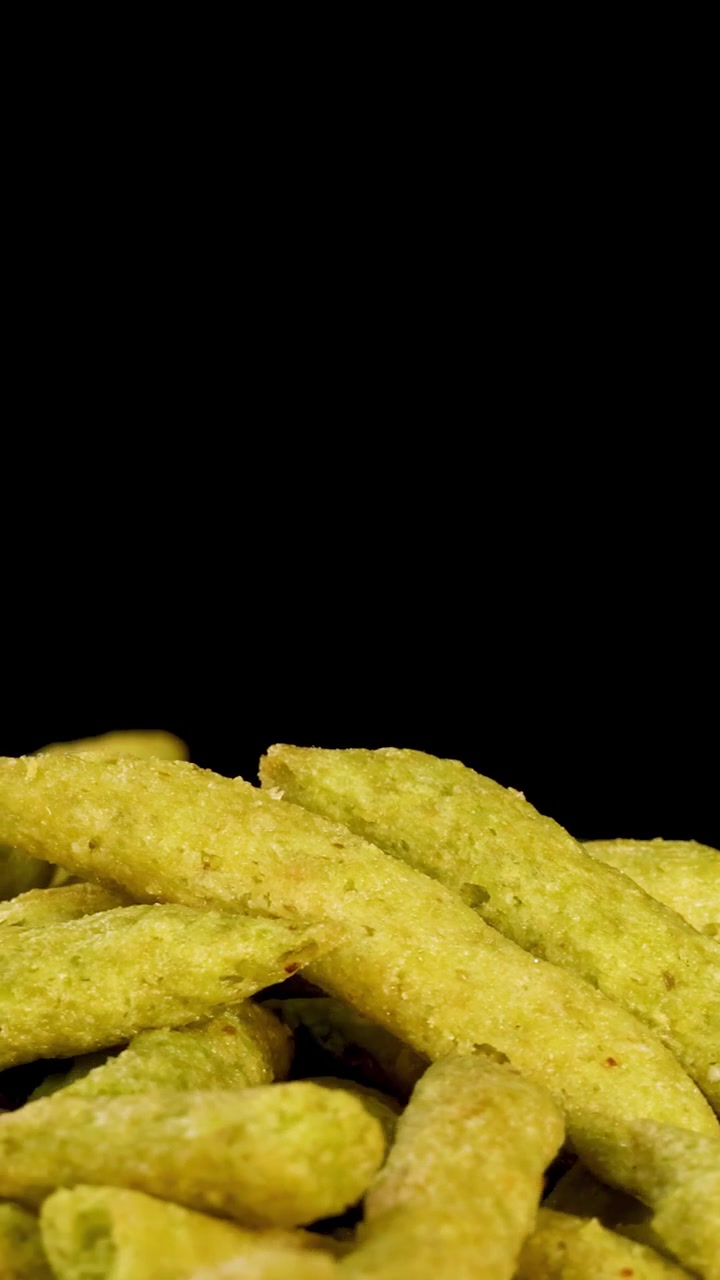 Una mano umana seleziona uno snack soffiato al pisello verde da una pila, sfondo nero, illuminazione da studio.