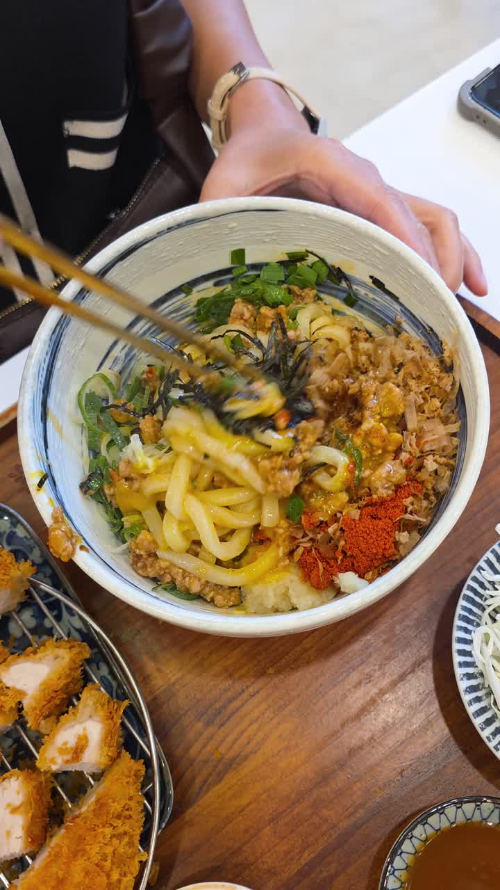 A close-up shows a person using chopsticks to mix a delicious bowl of Japanese Mazesoba, a noodle dish with minced pork, green onion, seaweed, fish roe, and a raw egg yolk