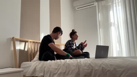 una pareja joven relajándose en una cama con computadoras portátiles y teléfonos