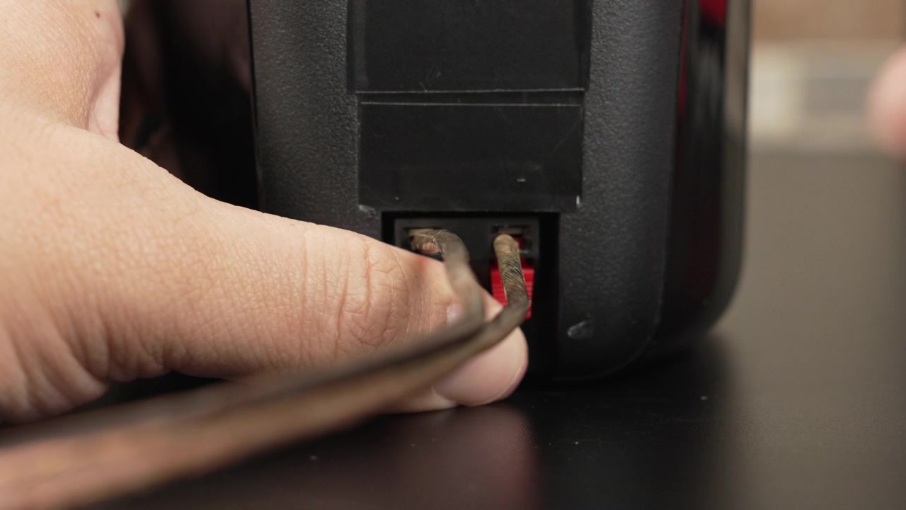 Hands attach bare speaker wire to the spring clip terminals on the back of a black bookshelf speaker on a desk, useful for tutorials, product demos, or home o guides.