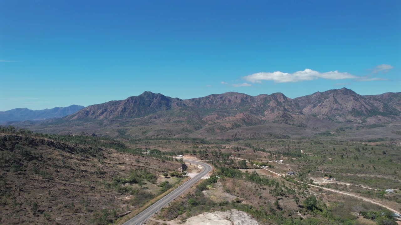 Ripresa cinematografica da drone ad alta quota che cattura un'autostrada a più corsie che attraversa un'ampia valle montana in un paesaggio remoto del Centro America. La scena presenta terreno di foresta secca, cieli blu e