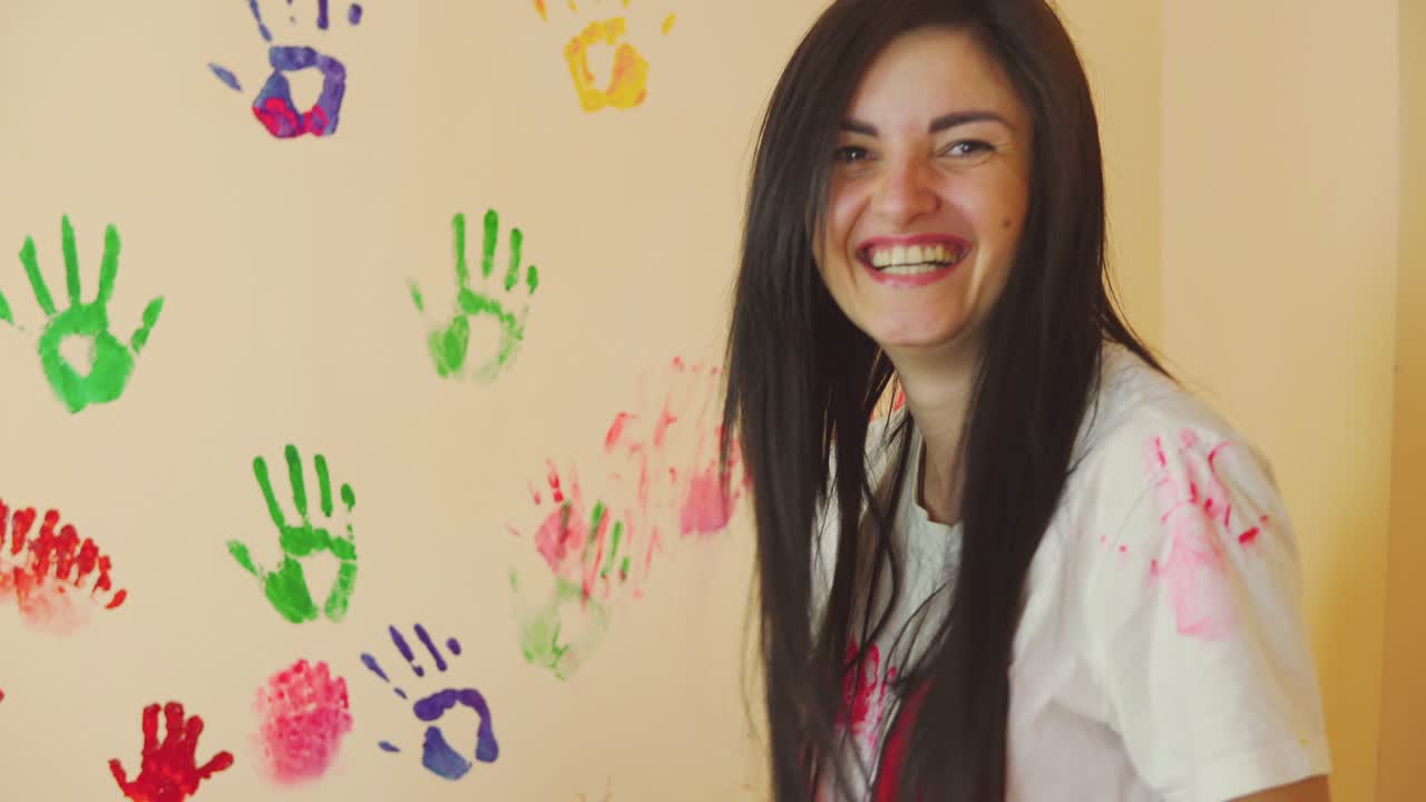 Happy mother and her cute boy having fun together leaving their colorful handprints on the wall and touching with painted hands each other. Mother leaves a painted spot on her son's nose.