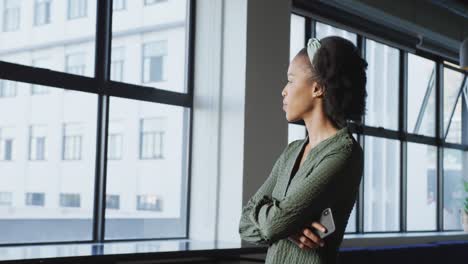 Happy-african-american-businesswoman-at-window-using-smartphone-alone-at-office