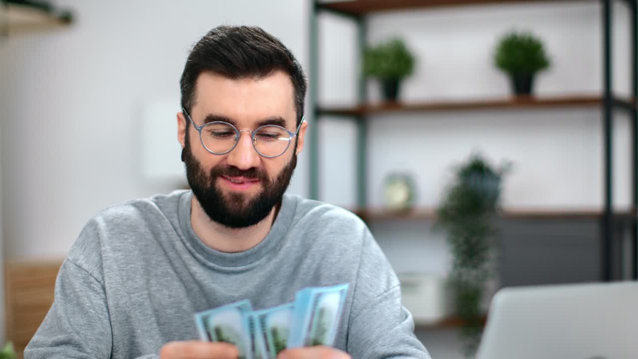 Hombre de negocios barbudo y feliz con gafas contando billetes de dólar en su lugar de trabajo