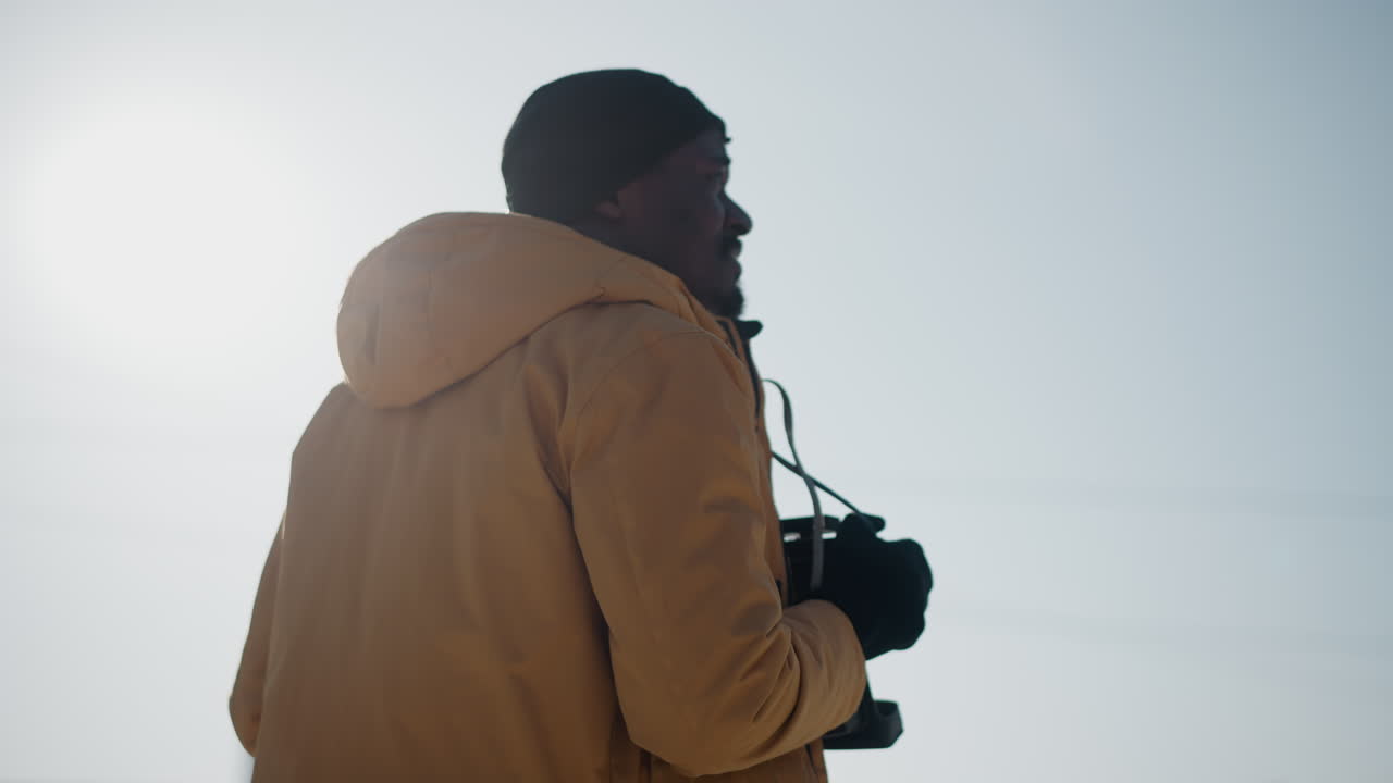 back view of snapper wearing beanie and winter jacket walking urban street with camera in hand, looking sideways under bright sun glare, breath in cold air, exploring snowy cityscape