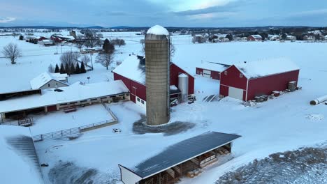 snowy farm with red barns, a silo, and bare trees under a cloudy sky