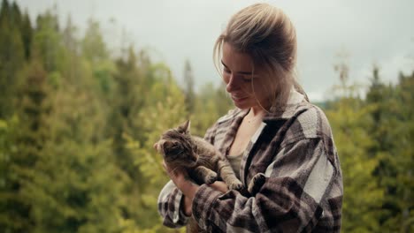 close-up shot: a happy blonde girl holds her cat in her arms and strokes her on the balcony against the backdrop of a coniferous forest in the mountains