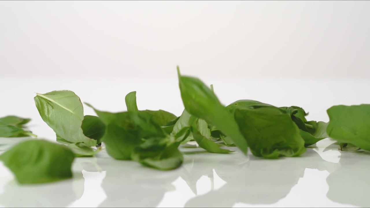 Foglie di basilico fresche e aromatiche che rimbalzano su un tavolo bianco in slow motion, in primo piano macro, prima di posarsi.