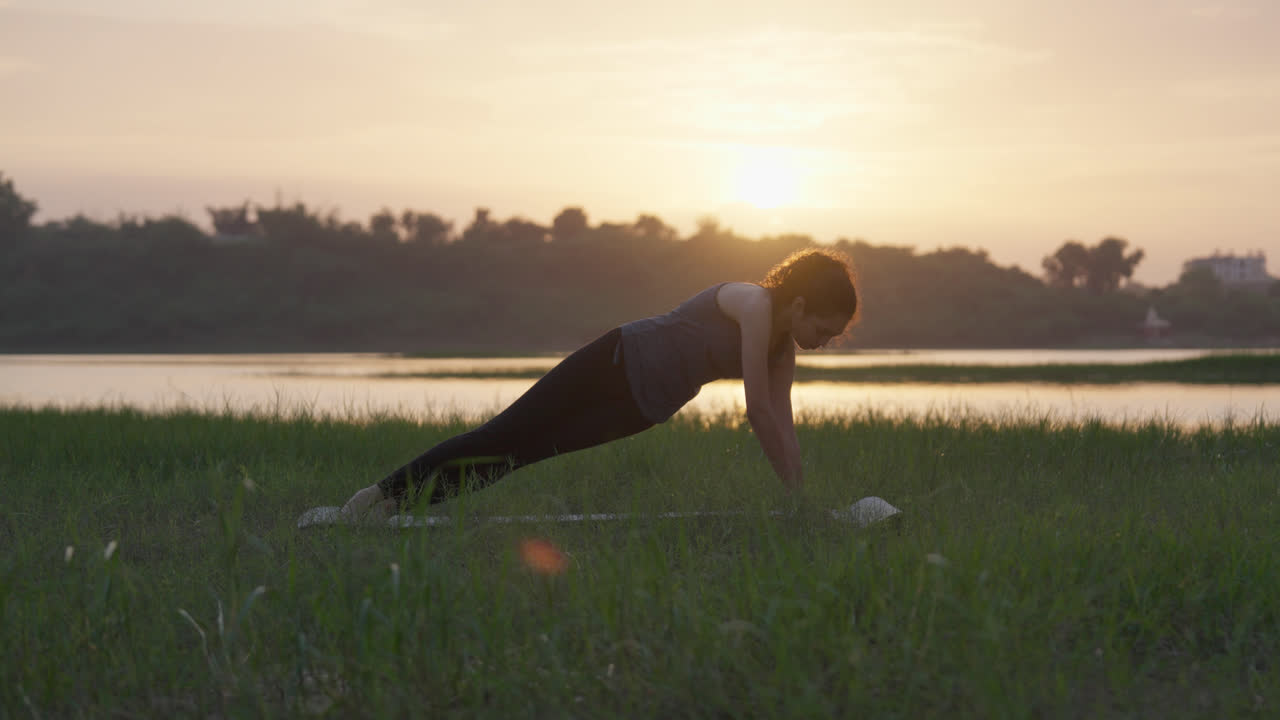 A woman practicing side plank yoga pose on grass during sunrise near a calm river landscape