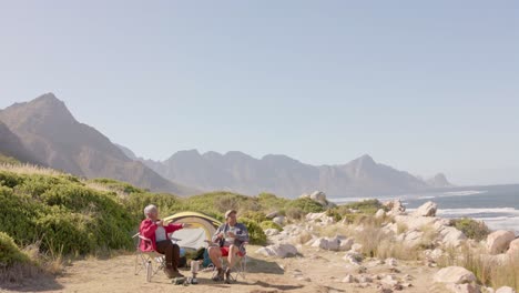 Happy-senior-biracial-couple-sitting-at-tent-in-mountains-and-drinking-coffee,-in-slow-motion