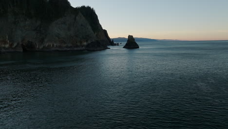 aerial dolly to sea stack rocks on the oregon coast with dusk glow of light yellow on horizon below exposed sea cliffs with tidal marks