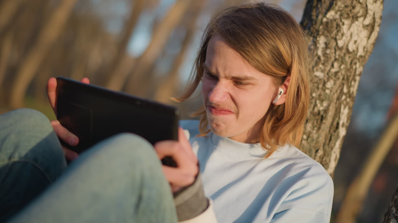young white teenager outdoors using tablet by tree, earphones in and concentrated expression while scrolling through content, casual jeans and warm sunlight filtering through bare branches, relaxed