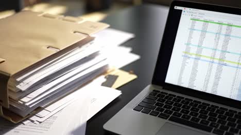 an organized workspace featuring a stack of documents next to a laptop displaying data on a spreadsheet, reflecting the diligence of administrative tasks
