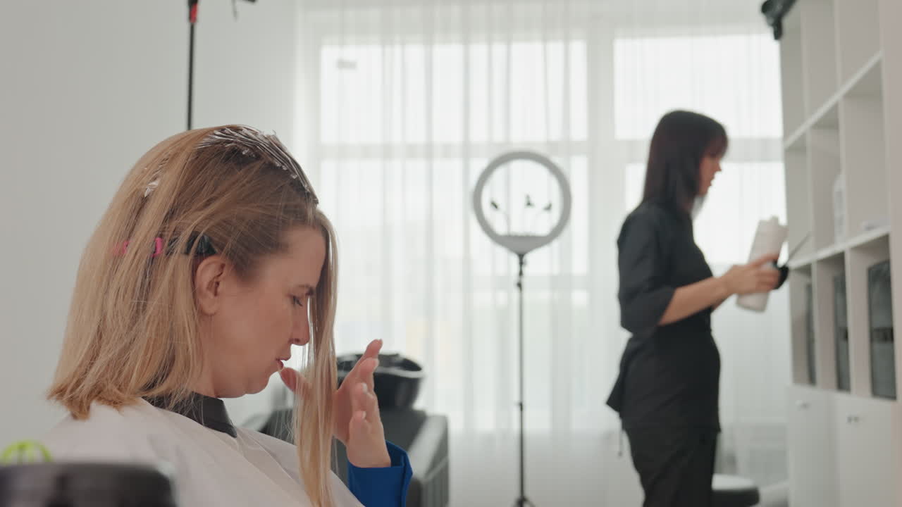 Nainen kampaamotuolissa laitattamassa raitoja, stylisti valmistelee tuotteita ja tarkistaa kaappia, rengasvalo ja ikkunan taustavalo, folioiden ja vaalennusaineen levitys, asiakas tutkii osioitua suortuvaa stylistin kanssa