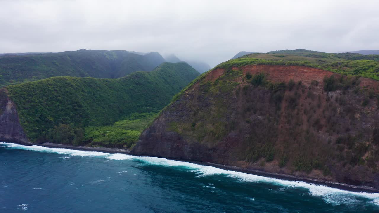 Sheer green cliffs of Polulu Valley, Hawaii extend into the Pacific Ocean, where vibrant blue waves crash powerfully along the dramatic volcanic coastline under cloudy skies.