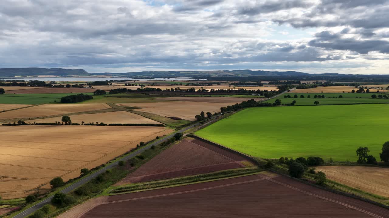 Drone footage glides above patchwork farmland with green, brown, and golden fields under cloudy skies. Smooth forward camera movement, wide landscape perspective, natural daylight
