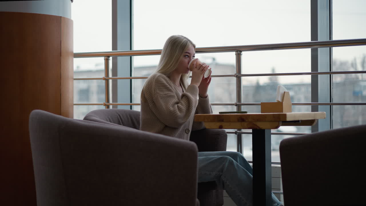 Sidebillede af kvinde, der nipper til kaffe med hånden hvilende på et træbord i et indkøbscenter, nyder sin drik, mens hun observerer omgivelserne gennem et vindue i et afslappet indendørs miljø.