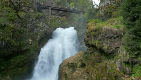 Suma-De-Cascada-Que-Cae-En-Cascada-En-Medio-De-Una-Vegetación-Verde,-Con-Un-Rústico-Puente-De-Madera-Arqueándose-En-Lo-Alto.