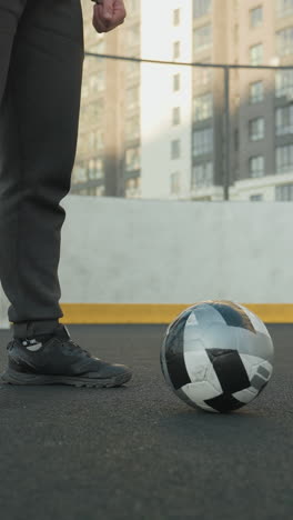 vista de ángulo inferior de un hombre en posición de cuclillas con las manos juntas, realizando un entrenamiento intenso en un campo deportivo al aire libre con una pelota de fútbol rodando en el suelo y un edificio urbano en el fondo