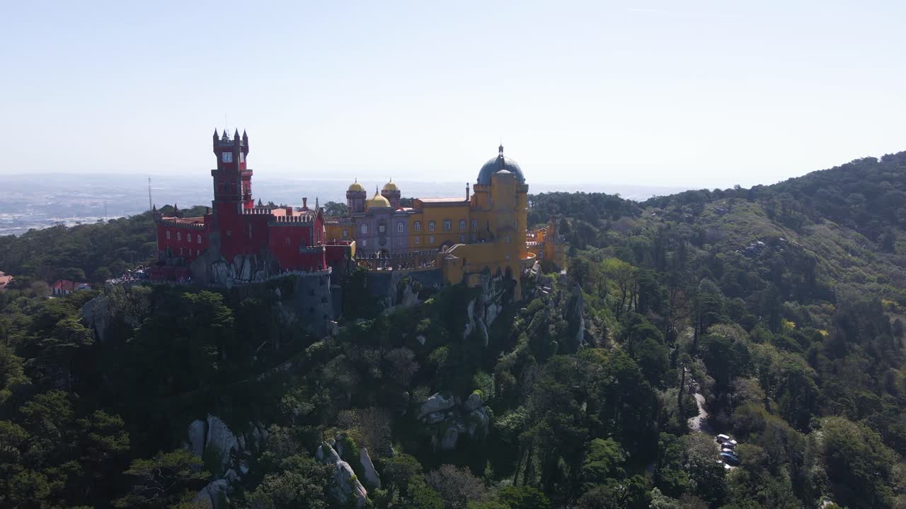 Luchtopname met drone van het iconische Pena Paleis in Sintra, met de kenmerkende rode toren, gele paleismuren, omgeven door weelderig bos en een Portugese stad zichtbaar in de vallei beneden.