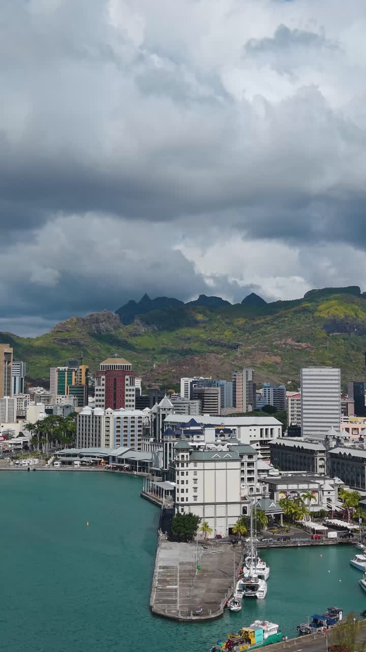 Vertical revealing aerial shot capturing the modern Port Louis skyline and the Caudan Waterfront set against the dramatic, rugged Moka Mountains of Mauritius. City meets nature