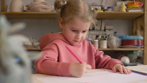 niña rubia dibujando en un papel sentado en una mesa en un taller de artesanía