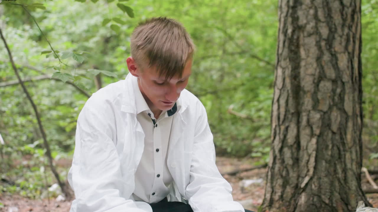 Close view of young man in white coat sitting on dry stump in forest removing glasses while showing fatigue and exhaustion during outdoor work surrounded by trees with bag placed beside him
