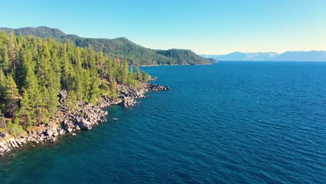 aerial drone flying over clear blue waters of lake george, new york with rocky shoreline and thick pine tree forest in mountains