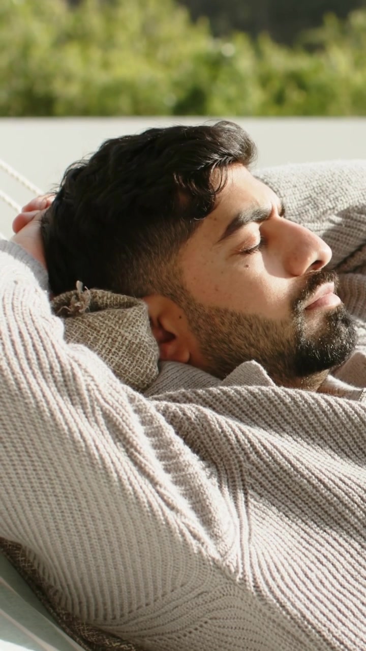 Video verticale: Uomo adulto che si crogiola al sole, si muove su un cuscino da patio mentre il sole lo illumina, indossando un maglione di lana.
