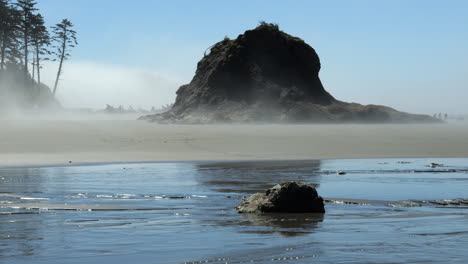 Niebla-Que-Sopla-A-Través-De-La-Playa-Con-Una-Gran-Formación-Rocosa,-Arena,-árboles-Y-Agua-Con-Reflejo