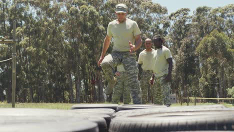 Eine-Vielfältige-Gruppe-Von-Soldaten-Beobachtet,-Wie-Ein-Männlicher-Ausbilder-Auf-Einem-Hindernisparcours-In-Der-Sonne-Durch-Reifen-Rennt