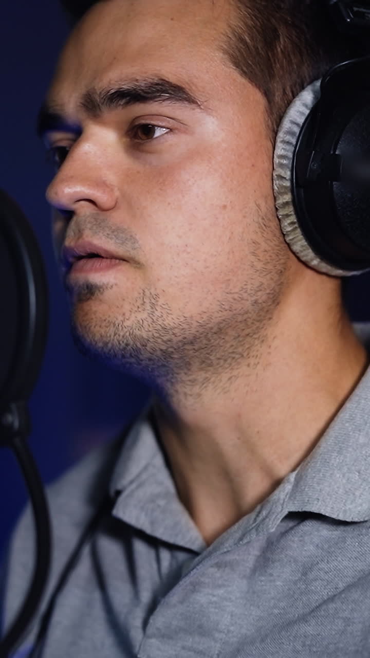 Singer recording a song in the studio. Male performer standing in front of the professional microphone and wearing headphones. Vertical video
