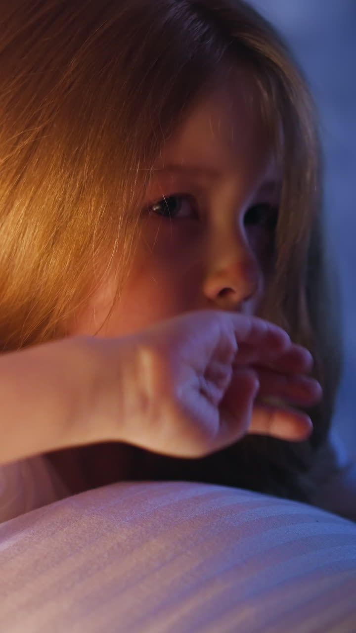 Upset blonde little girl with dimly lit face leaning on soft pillow wipes running nose and tears on bed near wall in semidark bedroom at moon light slow motion