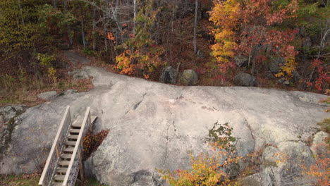 Aerial-drone-shot-rising-up-from-the-rock-face-of-a-dry-riverbed,-passing-over-forest-trees-revealing-the-beautiful-autumn-colors-as-nature-changes-between-seasons