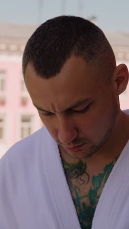 thoughtful young man with colorful tattoos on chest in white bathrobe smokes cigarette standing on terrace of hotel closeup slow motion
